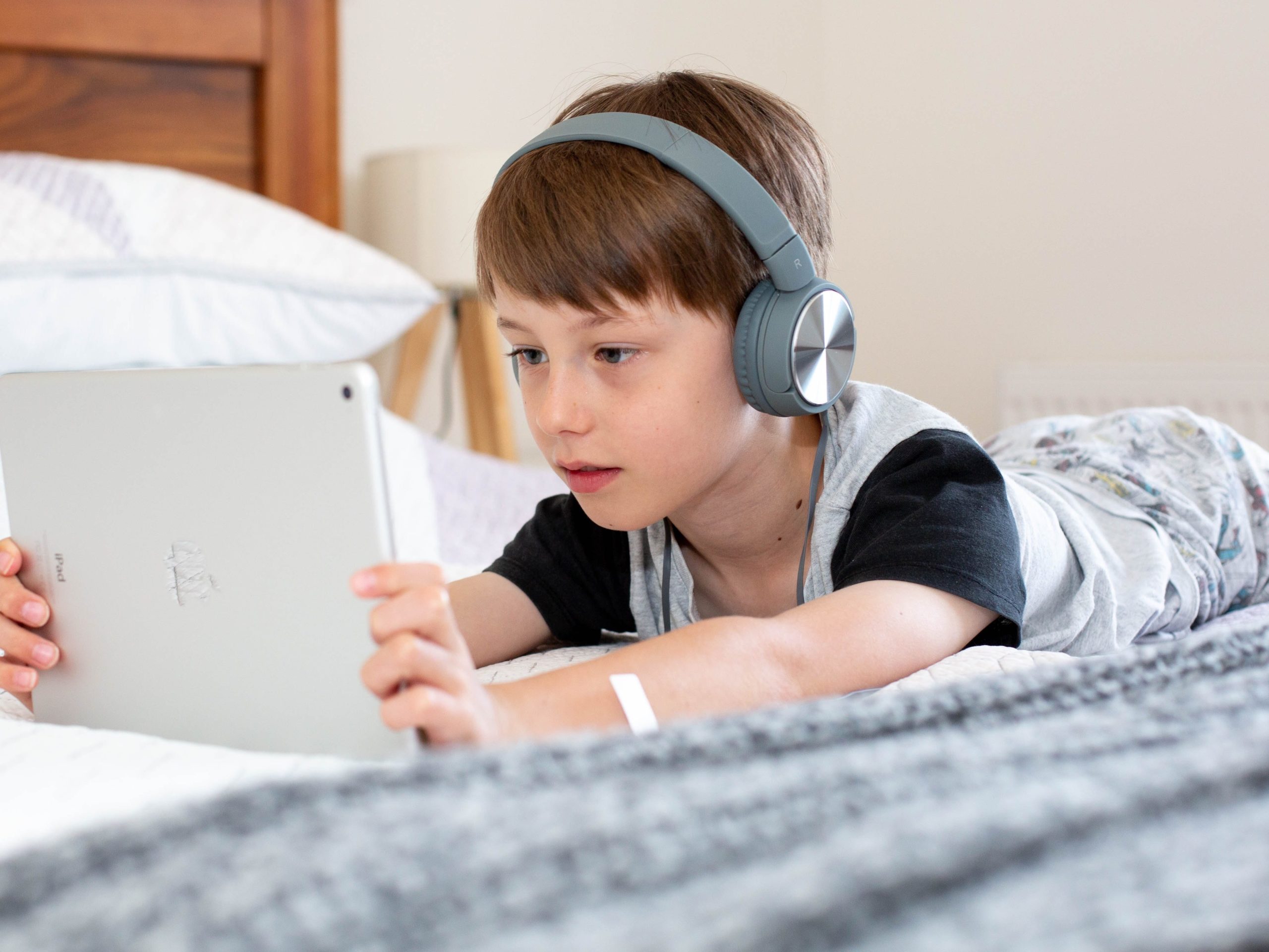 a boy in a grey and black shirt laying on the bed with grey headphones and watching something on an ipad. caribu mommy mom mother's blog that gives parenting advice for kids, children, toddlers, preschoolers.